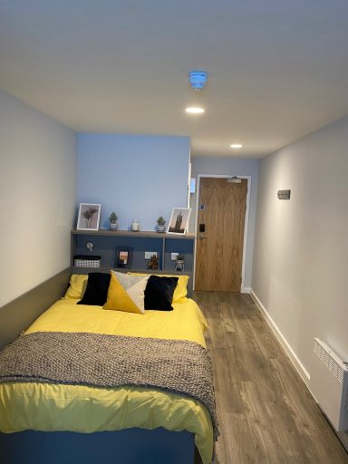 A modern bedroom with a yellow bedspread, shelves, and wooden door in a light-coloured room.